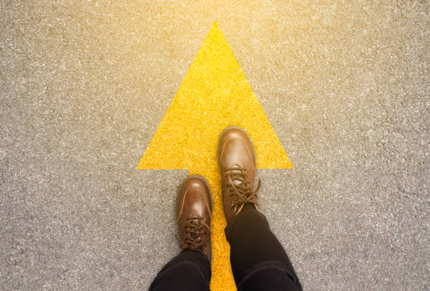 feet and arrows on road background in starting line beginning idea. top view. woman in leather ankle boots on pathway with yellow direction arrow symbol. moving forward, new start and success concept. - future imagens e fotografias de stock