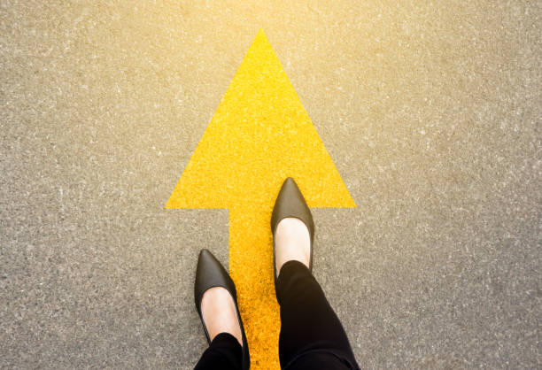 Feet and arrows on road background in starting line beginning idea. Top view. Business woman in black shoes on pathway with yellow direction arrow symbol. Moving forward, new start and success. Feet and arrows on road background in starting line beginning idea. Top view. Business woman in black shoes on pathway with yellow direction arrow symbol. Moving forward, new start and success. start over stock pictures, royalty-free photos & images