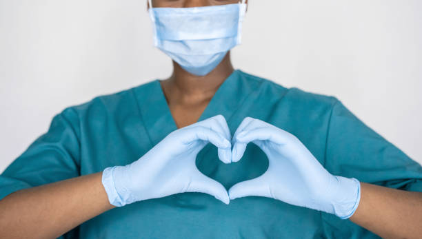 여성 아프리카 전문 의료진은 얼굴 마스크, 장갑, 심장 손 모양을 보여주는 파란색 녹색 유니폼을 착용합니다. 의료 사랑, 관리 및 안전 기호, 코로나 바이러스 건강 보호 기호 개념. 근접 촬영 - nurse 뉴스 사진 이미지