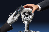女性のAIロボットビジネスマンは、スマートフォンでロボットの頭蓋骨を制御します。人間の手が金属の頭蓋骨を握っている。