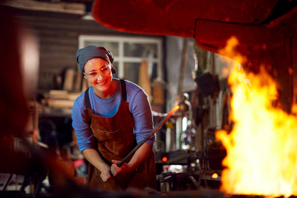 Female Blacksmith Heating Metalwork In Flames Of Forge Female Blacksmith Heating Metalwork In Flames Of Forge blacksmith stock pictures, royalty-free photos & images