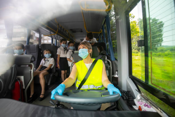 COVID-19. Female bus driver transporting elementary school kids by bus. Young female bus driver driving he school bus, wearing a face mask. She is taking care that kids sit down. school bus driver stock pictures, royalty-free photos & images