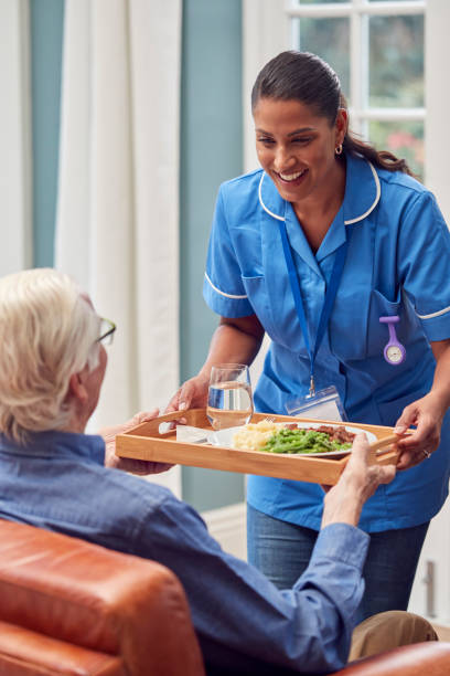 Female Care Worker In Uniform Bringing Meal On Tray To Senior Man Sitting In Lounge At Home stock photo