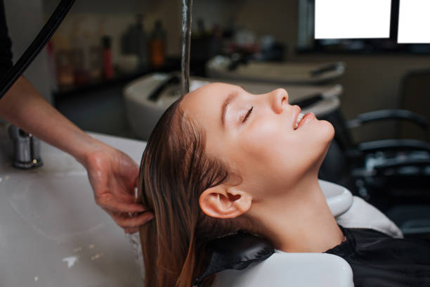 Female client in beauty salon. Side view of beautiful young woman with closed eyes smiling while hairdresser rising hair after shampoo, hair spa stock photo