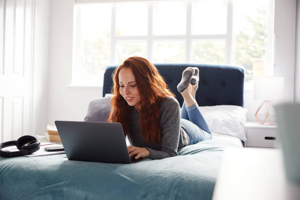 Female College Student Lies On Bed In Shared House Working On Laptop Female College Student Lies On Bed In Shared House Working On Laptop college dorm stock pictures, royalty-free photos & images