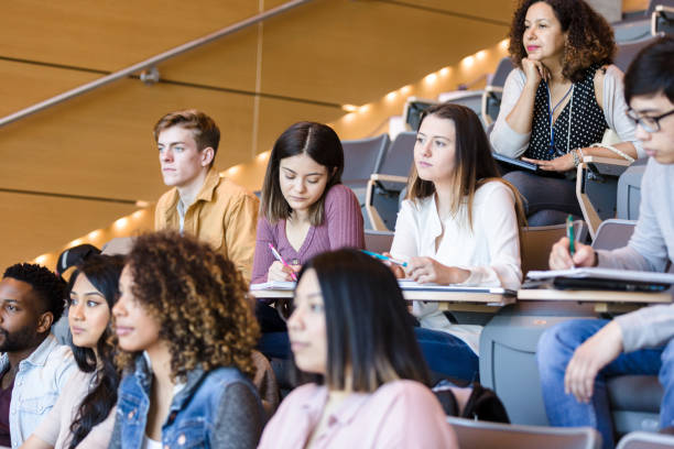 Female college student taking notes A young Hispanic female college student takes notes during class. Other students are attentively listening to a lecture. community colleges stock pictures, royalty-free photos & images