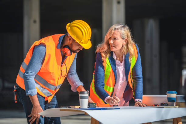 Mature female construction site coordinator standing at planning table with young African foreman and discussing details.