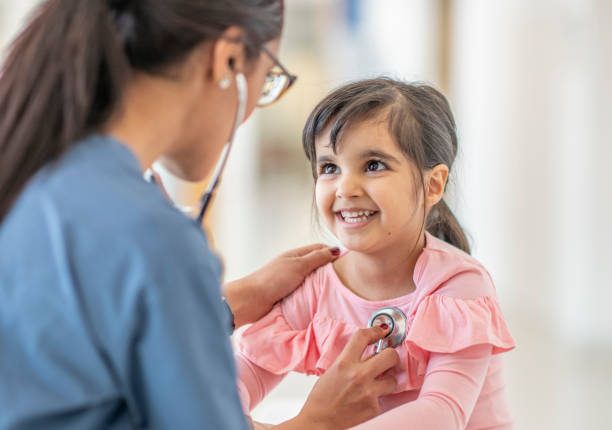 Female doctor checks girl's heartbeat A toddler girl sits on a table during a medical examination. A female medical professional is using a stethoscope to listen to her heartbeat. The child is smiling up at her doctor. pediatrician stock pictures, royalty-free photos & images