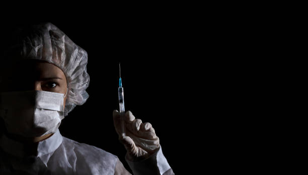 Female doctor in protection clothing holds syringe with vaccine in her hand on black background. Half face in shadow, dramatic emotion. Concept of virus protection, prevention of epidemics, treatment of diseases Female doctor (or nurse) in protection face mask, medical cap and glove holds syringe with vaccine in her hand on black background. Half face in shadow, dramatic emotion. Concept of virus protection, prevention of epidemics, treatment of diseases. Banner, copy space dose photos stock pictures, royalty-free photos & images