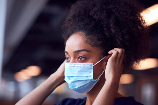 Female Doctor In Scrubs Putting On Face Mask Under Pressure In Busy Hospital During Health Pandemic Female Doctor In Scrubs Putting On Face Mask Under Pressure In Busy Hospital During Health Pandemic applying stock pictures, royalty-free photos & images