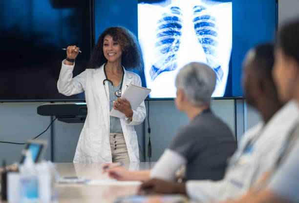 Female Doctor Leading a Meeting A female doctor of African decent stands at the front of a room as she leads a team meeting to discuss a patients plan of care.  Her colleagues are seated at a large table in front of her as she talks about the x-rays displayed on the screen behind her.  They are each dressed professionally in scrubs and lab coats. best medical schools stock pictures, royalty-free photos & images