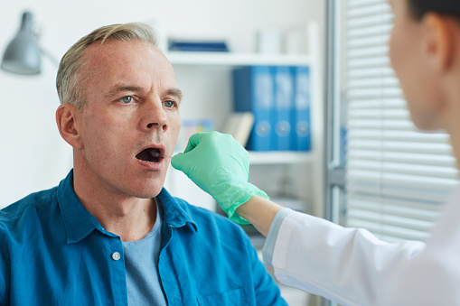 Unrecognizable female doctor taking swab sample from mouth and throat of mature man for virus test in clinic, copy space