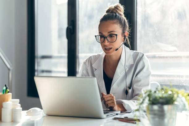 Female doctor talking while explaining medical treatment to patient through a video call with laptop and earphones in the consultation. stock photo