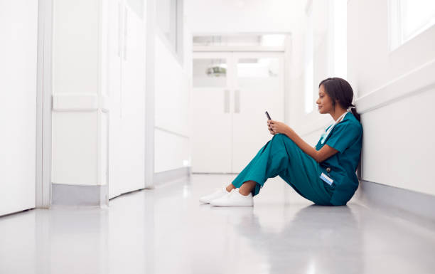 Female Doctor Wearing Scrubs Sitting On Floor In Hospital Corridor Using Mobile Phone Female Doctor Wearing Scrubs Sitting On Floor In Hospital Corridor Using Mobile Phone fomo stock pictures, royalty-free photos & images
