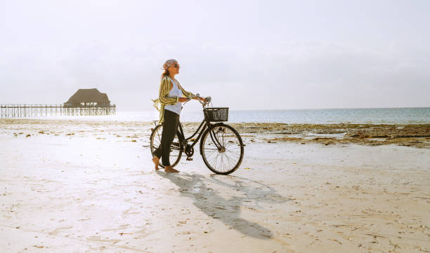 Female dressed light summer clothes have morning walk with old vintage bicycle with front basket on the lonely low tide ocean white sand coast on Kiwengwa beach on Zanzibar island, Tanzania. stock photo