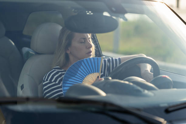 Female driver with hand fan suffering from heat in car, has problem with non-working air conditioner stock photo