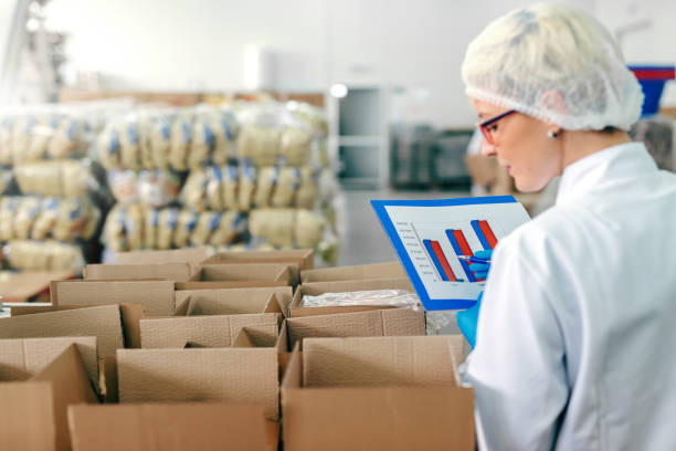 female employee in sterile uniform and with chart in hands counting products in boxed. food factory interior. - food chart healthy imagens e fotografias de stock