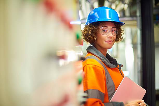 female engineer A female industrial service engineer has just conducted a safety check of a control panel in a boiler room. She is wearing hi vis, hard hat, safety glasses and holding a digital tablet that she used to conduct a safety inspection. The engineer is stated next to the control panel looking to camera and smiling. hardhat stock pictures, royalty-free photos & images
