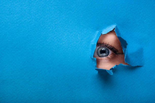 A female eye looks through a hole in a paper blue background. stock photo