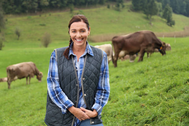 Female farmer portrait on pasture Mid-adult woman standing on a grassland with group of cows behind her. rancher stock pictures, royalty-free photos & images