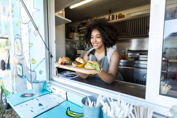 Female food vendor offering sandwiches in food van Smiling female food vendor offering sandwiches and burgers in food van. food truck stock pictures, royalty-free photos & images
