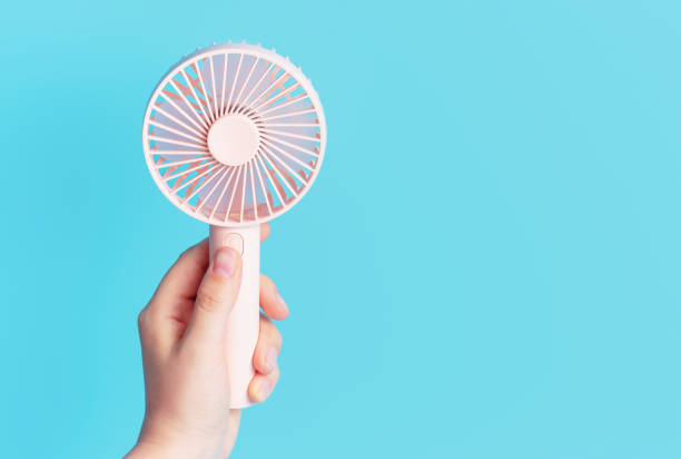 Female hand holding a pink mini fan on blue background close-up. stock photo