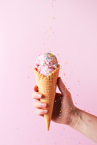 Female hand holding the pink ice cream with falling sprinkles in waffle cone on pink background