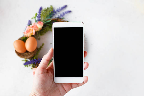 Female hand with a phone takes pictures of an Easter wreath with bright spring flowers, a nest with Easter eggs stock photo