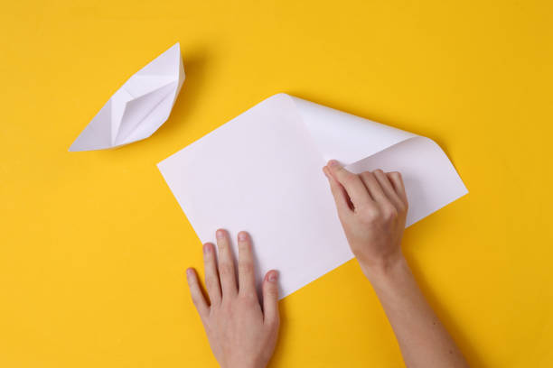 Female hands fold origami boat on yellow background. Top view stock photo