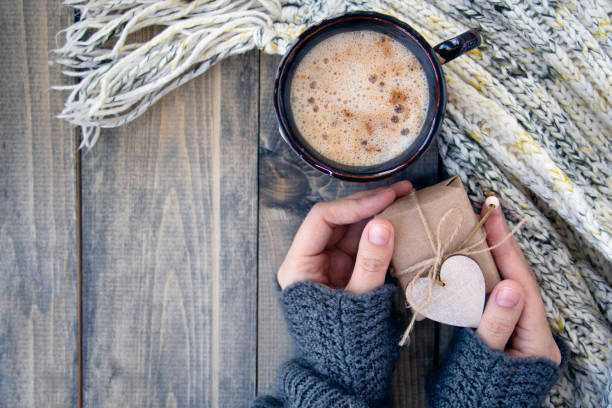 Female hands hold little gift near cup with a hot drink on of wooden table with warm knit woolen scarf. Christmas background - The concept of winter, warmth, holidays and events.