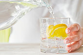 istock Female hands pouring water from the decanter into a glass beaker with lemon and ice. Health and diet concept. Quenching thirst on a hot day. 1309474558