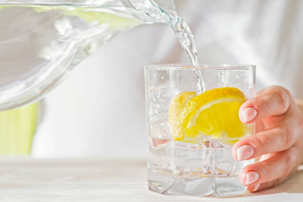 mani femminili che versano acqua dal decanter in un bicchiere con limone e ghiaccio. concetto di salute e dieta. dissetarsi in una giornata calda. - bere acqua foto e immagini stock