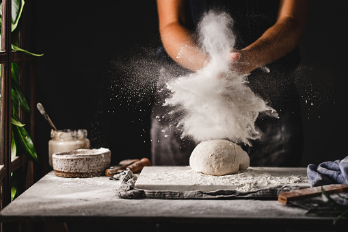 Midsection of a woman throwing flour on dough over marble board. Female preparing sourdough bread in the kitchen.