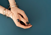 istock Female hands with trendy dark nail design with gold bracelets on aqua background. Luxury concept. Festive backdrop for your design. 1299139185