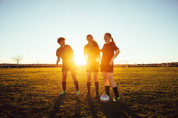 Female High School Soccer Players Three female high school soccer players in practice uniform pause for a candid moment while on the pitch in Arizona, USA. The three friends and teammates enjoy their friendship at dusk with the sun shining through their silhouettes. high school sports stock pictures, royalty-free photos & images