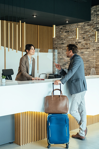 Business man with luggage talking with concierge on hotel reception with sneeze guard protection