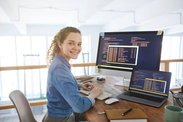 Female IT Developer at Workplace Portrait of female IT developer smiling at camera while typing on keyboard with black and orange programming code on computer screen and laptop in contemporary office interior, copy space computer programmer photos stock pictures, royalty-free photos & images