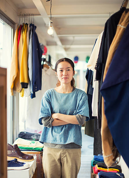 Female Japanese Resale Shop Owner in her Store A Japanese female owner of a second-hand clothing shop poses for a portrait in Tokyo, Japan. She is confidently standing with arms folded in the window light of her business. She is smiling and excited for business while looking at the camera surrounded by clothes for sale. thrift store photos stock pictures, royalty-free photos & images