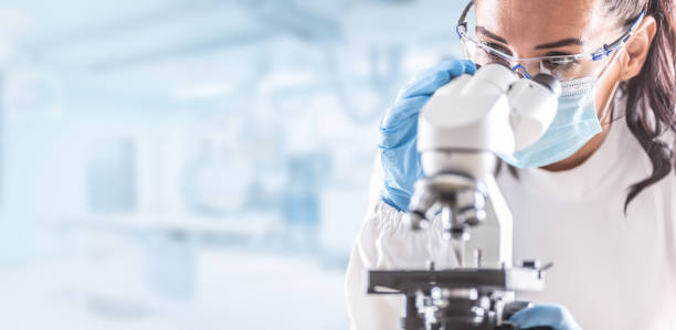female lab technician in protective glasses, gloves and face mask sits next to a microscope in laboratory. - investigação assunto imagens e fotografias de stock