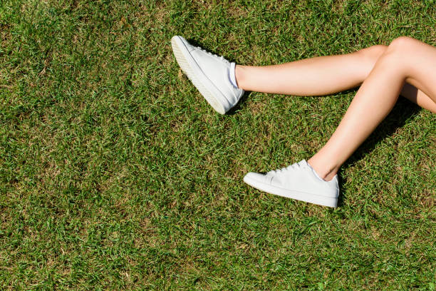 le gambe femminili in scarpe da ginnastica bianche giacciono sull'erba in un parco. - shoes top view foto e immagini stock