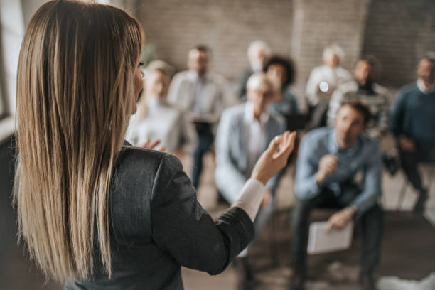Female manager talking to large group of her colleagues on a business seminar. Back view of female public speaker giving a presentation to large group of business people in a board room. public speaking stock pictures, royalty-free photos & images