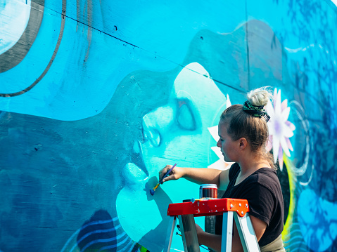 Female mural artist creating art on the wall exterior in public park in the city.