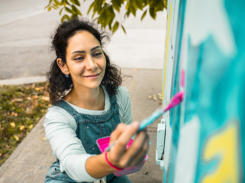 Female mural artist creating art on the public utility box on the street corner in the city.