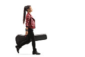 Full length profile shot of a female musician walking and carrying a guitar in a case isolated on white background