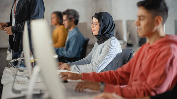 female muslim student in hijab, studying in university. she works on desktop computer in college with diverse multiethnic classmates. applying her knowledge to acquire academic skills in class. - arabic student stockfoto's en -beelden