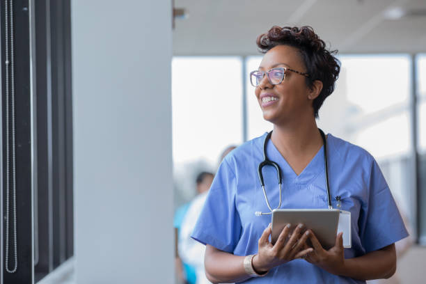 Female nurse holding digital tablet smiles while looking out window Female nurse holding digital tablet smiles while looking out window administrator stock pictures, royalty-free photos & images