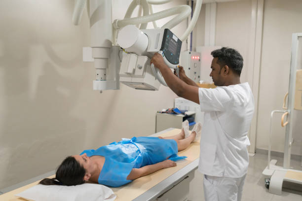 Female patient lying down ready for an xray and radiologist getting ready the machine Female patient lying down using a hospital gown ready for an xray and radiologist getting ready the machine x ray stock pictures, royalty-free photos & images
