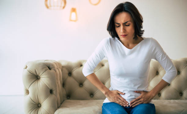 Female problems. Adult woman is sitting on a sofa at home and touching her lower stomach while suffering from cramps. Adult woman is sitting on a sofa at home and touching her lower stomach while suffering from cramps. endometriosis photos stock pictures, royalty-free photos & images