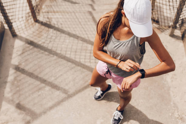 Female runner looking at smart watch heart rate monitor Young woman checking progress on smart watch. Female runner looking at smart watch heart rate monitor. cap hat stock pictures, royalty-free photos & images