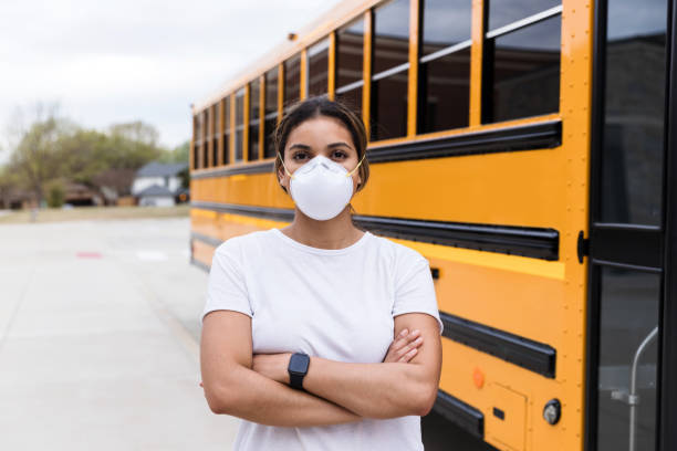Female school bus driver wearing N95 mask A female school bus driver, wearing an N95 mask, stands next to a school bus. She is standing with her arms crosses and is looking at the camera. school bus driver stock pictures, royalty-free photos & images